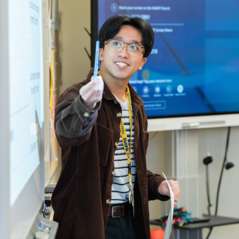 A man stands in front of a white board and points with a pen in hand.