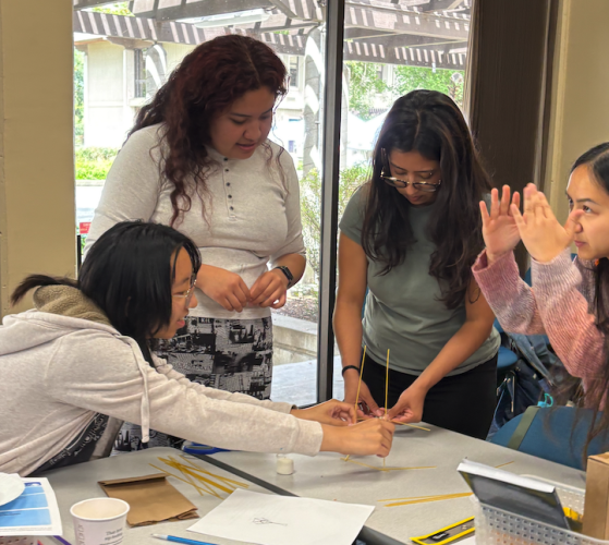 Four credential candidates stand over a table and collaborate on a hands-on project in front of them.