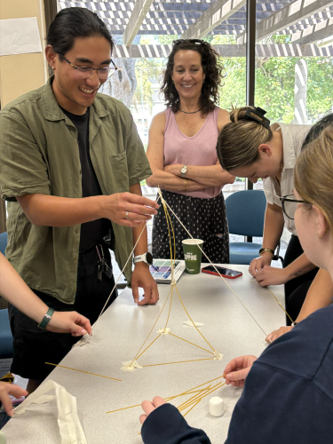 A man wearing a green shirt stands at a table and forms raw noodles into a structure. A woman wearing a pink shirt and a group of other participants stand around him.