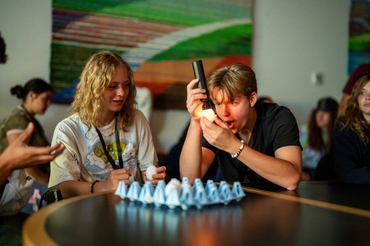 A high schooler shines a flashlight down on a chicken egg and smiles, while another student sitting next to him looks on.