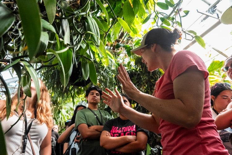A woman wearing a pink shirt and brown hat addresses a group of students standing around her in a greenhouse.