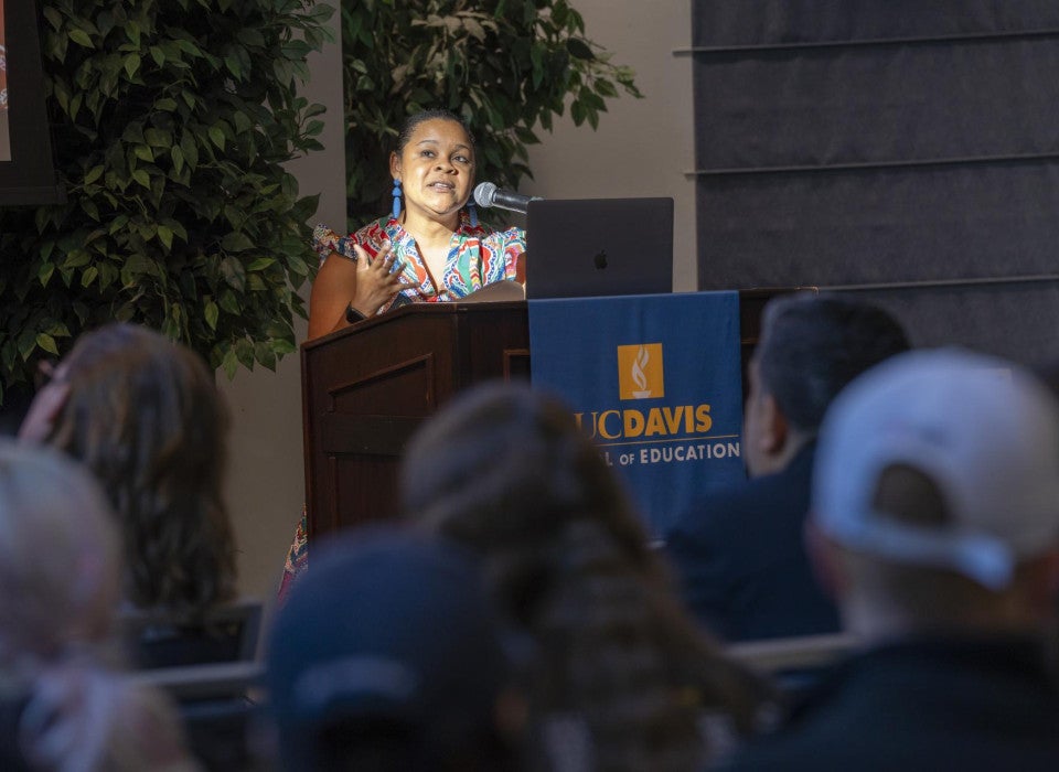 A woman stands at a podium with a banner that reads "UC Davis School of Education" and addresses a crowd of people sitting in front of her.