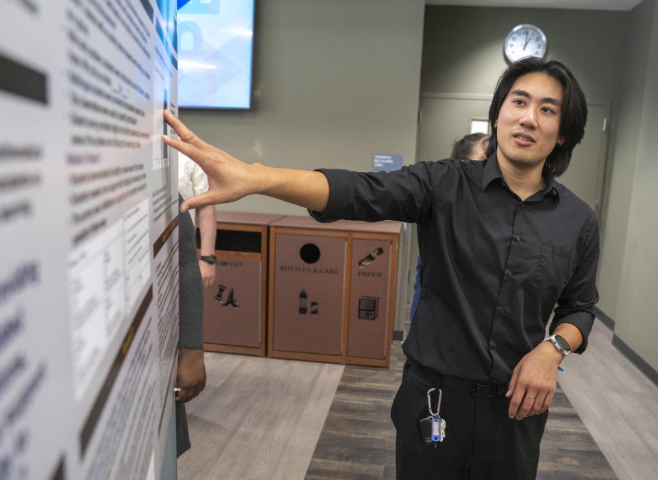 A man in a black shirt is presenting a large research poster, gesturing with one hand.