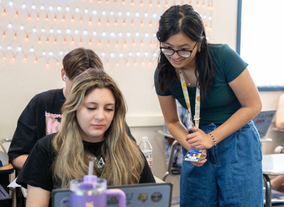 A teacher wearing a lanyard and glasses leans over a student's desk. They both look at a laptop screen together.