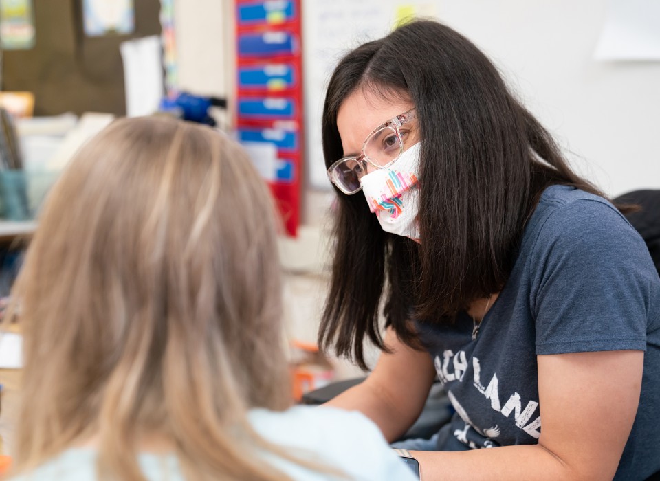 An elementary school teacher wearing a mask providing instructor to a student