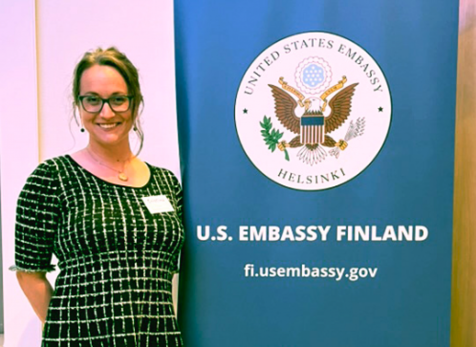 Cristina Buss stands next to a sign for the U.S. Embassy in Finland.