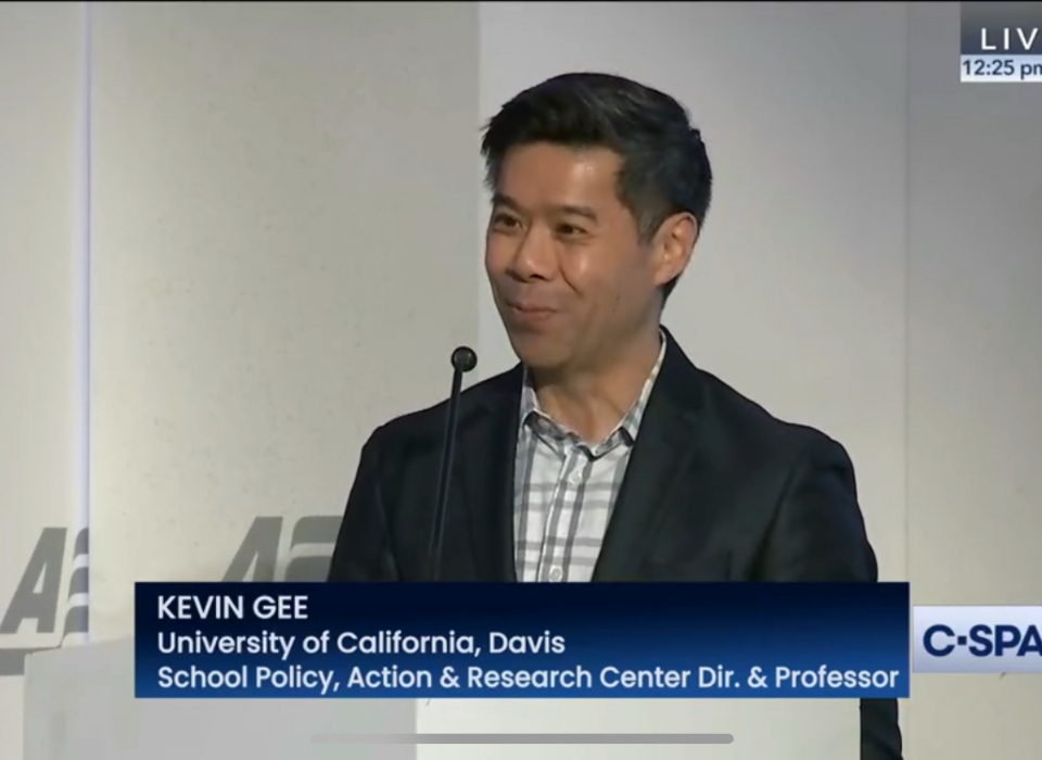 A video screenshot of Kevin Gee, standing at a podium and speaking into a microphone.