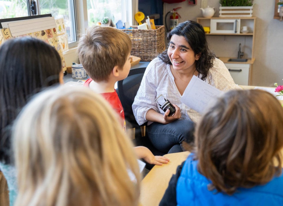 A woman wearing a white blouse and blue jeans sits at a table and smiles at four students standing in front of her.