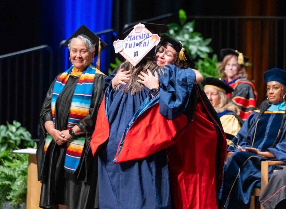 Two women wearing regalia embrace on stage during the graduation ceremony.