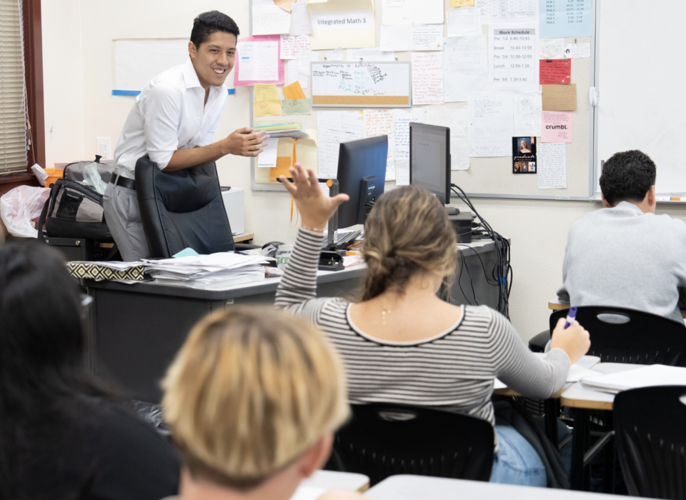 A girl raises her hand and addresses a teacher who is standing at a desk in front of her.