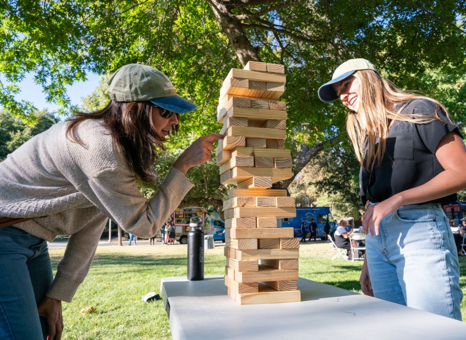 Two people - one wearing jeans, a pink sweater, and a green and blue School of Education hat and the other wearing jeans, a black shirt and the same hat - pull a block out of a Jenga tower.