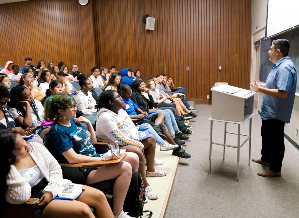 Students in a college classroom listen to a professor at a podium