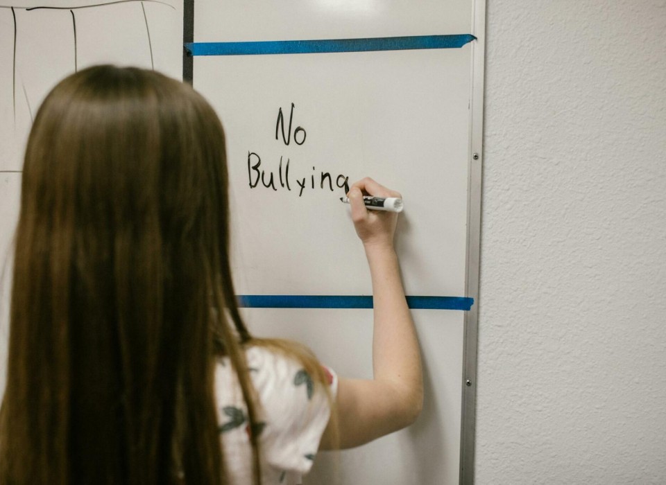 Young female student faces away from camera and writes "no bullying" on a white board in front of her.