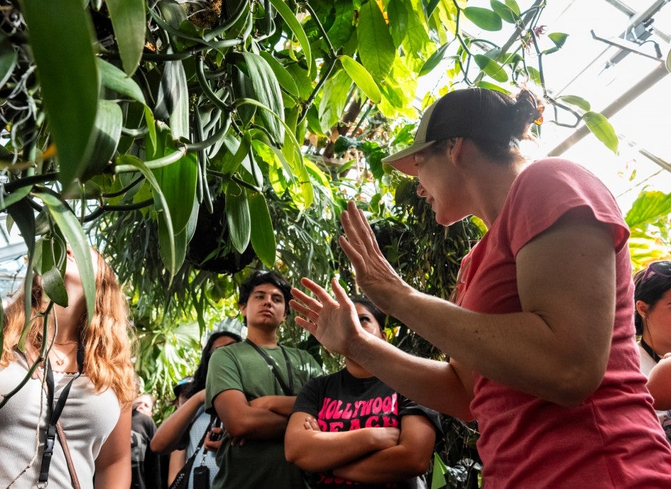 A woman wearing a pink shirt and brown hat addresses a group of students standing around her in a greenhouse.