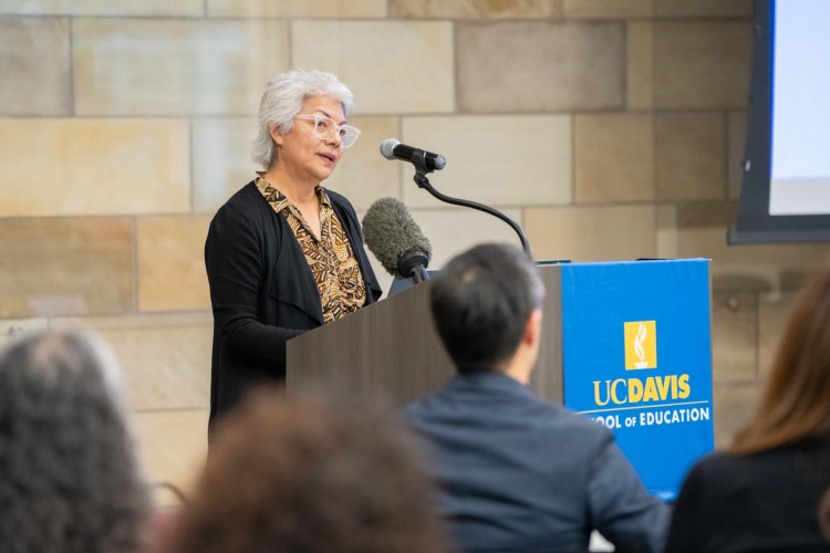 A woman stands at a podium in front of an audience, speaking into a microphone.