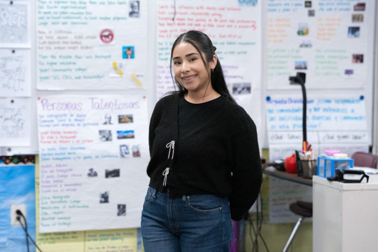 A bilingual teacher smiles in her classroom, with posters in Spanish and English behind her.