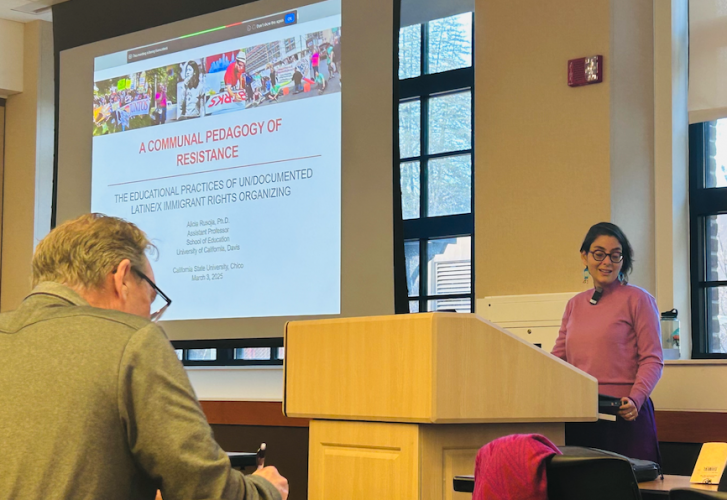 Alicia Rusoja stands at a podium, next to a projector screen that reads "A Communal Pedagogy of Resistance."