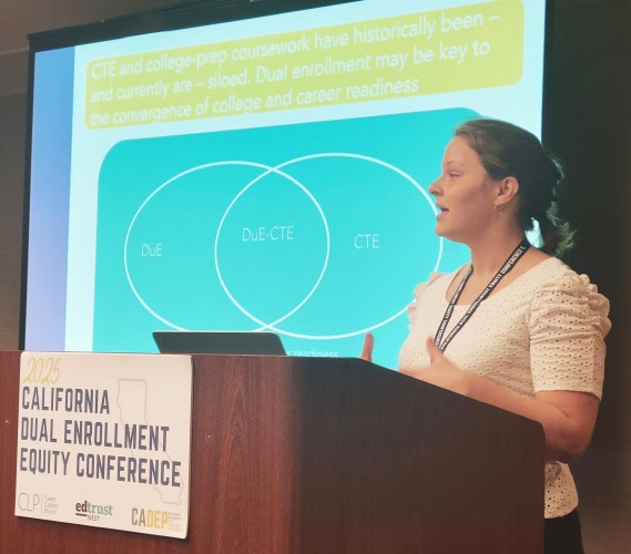 Audrey Boochever speaks at a podium. Behind her is a presentation screen with the words "CTE and college-prep coursework have historically been—and currently are—siloed."