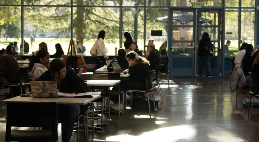 A student wearing headphones sits at a table by herself in a crowded dining hall.