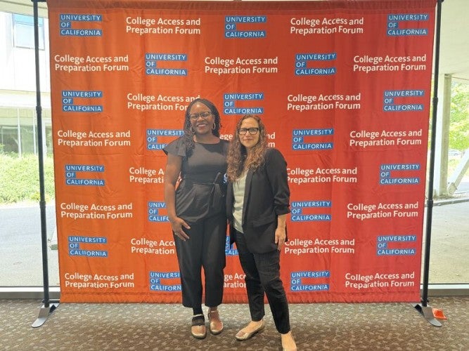 Two women wearing professional clothing stand together in front of a red photo background with "CAP Forum" printed on it.
