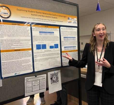 A woman wearing a suit stands in front of poster and gestures toward it with her hands.