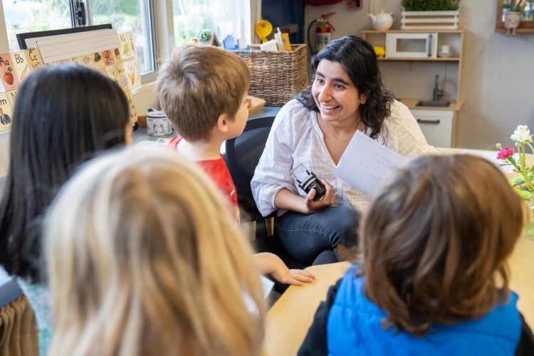 A woman wearing a white blouse and blue jeans sits at a table and smiles at four students standing in front of her.