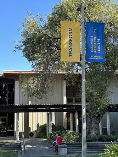 Photo of School of Education building showing two people sitting and chatting under banners which read  "Transform" and "Teachers Researchers Leaders"