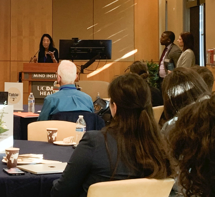 Nancy Tseng stands at a podium and speaks into the microphone. To her left stand Charles Wilkes and Nicole Sparapani, respectively