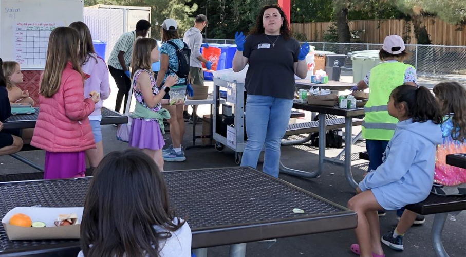 Students sit at a school lunch area while a woman wearing a navy shirt and jeans addresses them.