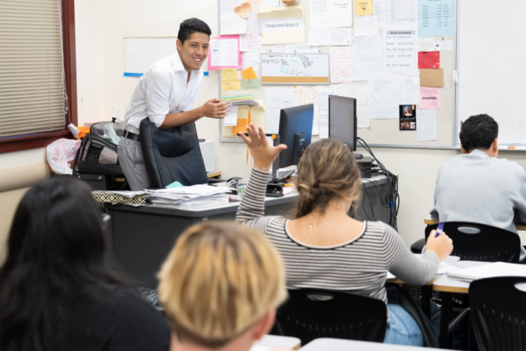 A student raises her hand and addresses a teacher who is standing in front of her, at a desk.
