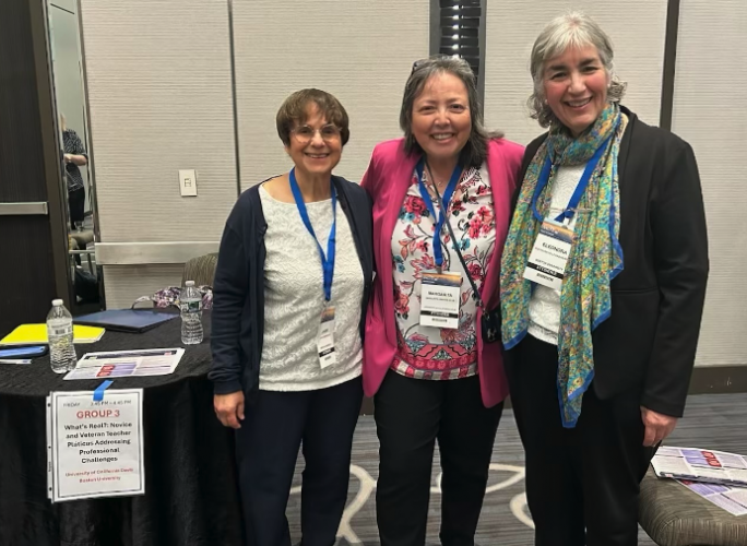 Three women pose together in front of a table with a sign that reads "'What's Real?': Novice and Veteran Teacher Pláticas Addressing Professional Challenges."