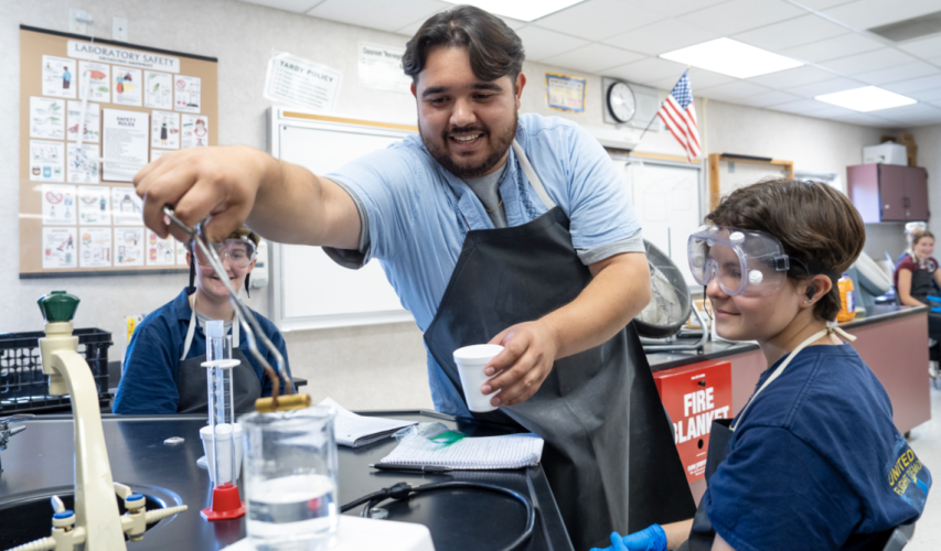 A man stands in a science classroom, wearing a protective apron. He places tongs into a glass beaker while two students wearing protective equipment watch.