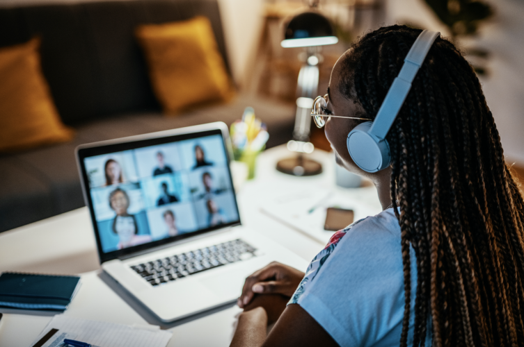 A woman wearing headphones looks at a laptop screen where nine people are speaking on a video conference.
