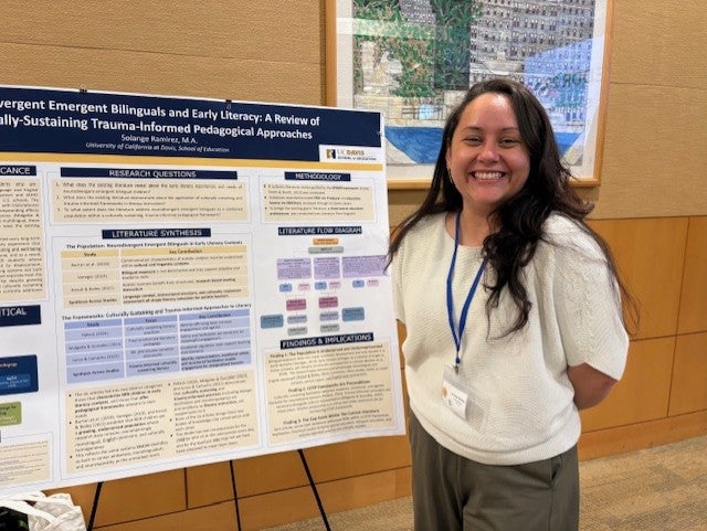 Solange Ramirez wears a white shirt and blue lanyard and stands in front of a research poster and smiles.