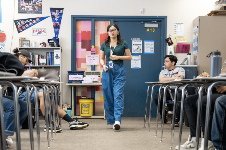 A woman wearing blue jeans, a green shirt, and a lanyard walks down an aisle between two rows of desks.