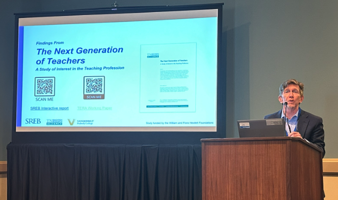 Tom Smith stands at a podium and speaks into a microphone. Next to him is a projector screen that says "The Next Generation of Teachers: A Study of Interest in the Teaching Profession."